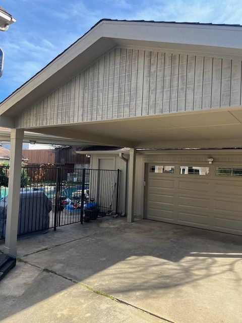 Carport beside a beige garage with a metal gate and trash bins, under a bright blue sky