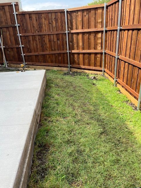 Backyard corner with grass strip beside a concrete patio and wooden privacy fence.