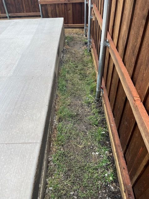Narrow strip of grass and dirt between a concrete patio and a wooden fence.