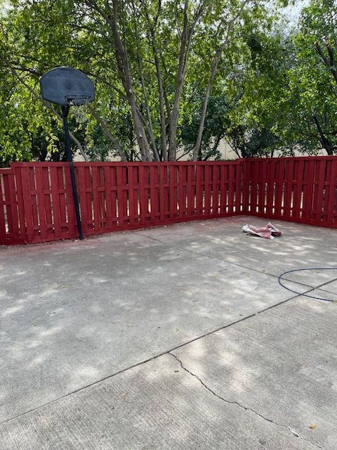 Concrete patio with a red fence, basketball hoop, and trees in the background.