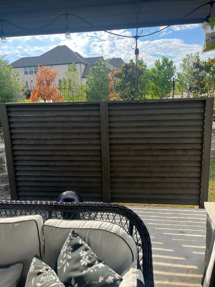 Patio seating facing a dark privacy fence, with trees and houses visible beyond in daylight.
