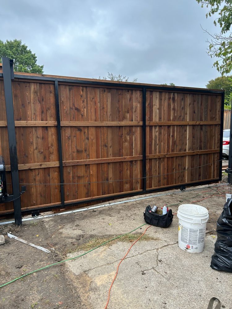 Wooden fence with black metal posts beside a driveway; bucket, shoes, and orange extension cord in foreground.