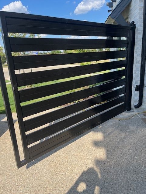 Black slatted metal driveway gate beside a concrete wall under a blue sky