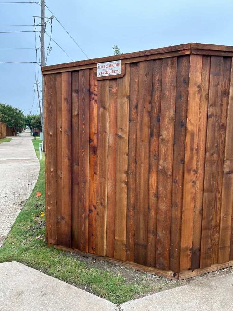 Wooden utility enclosure beside a sidewalk, with a small sign on the front and power lines overhead.