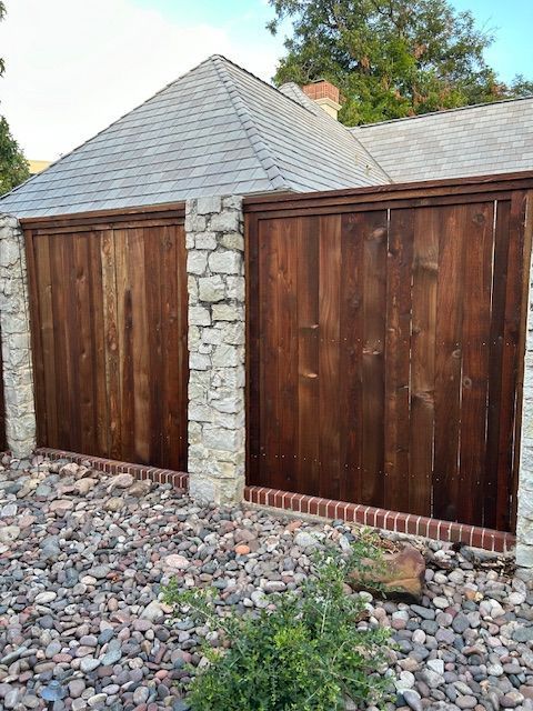 Stone-and-wood fence with two dark wooden gates beside a gravel yard and trees behind.