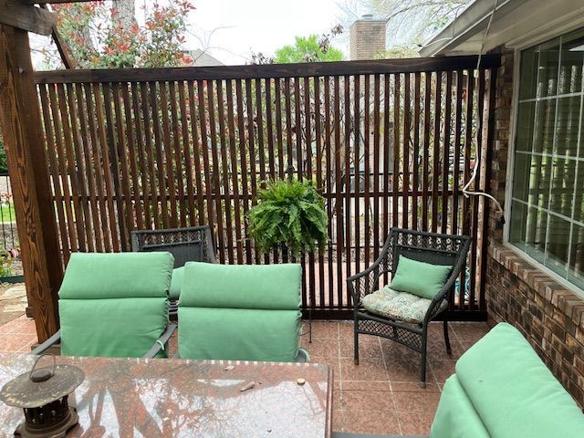 Covered patio with green cushioned chairs, a black chair, potted plant, and slatted wooden privacy screen.