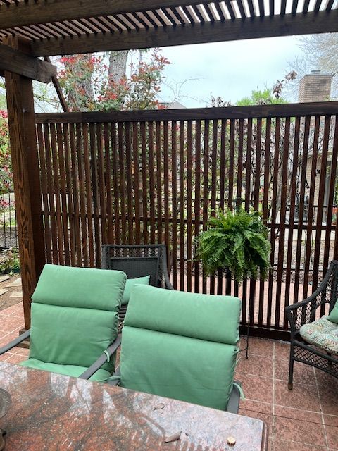 Outdoor patio with green-cushioned chairs, glass table, potted fern, and wooden privacy screen under a pergola