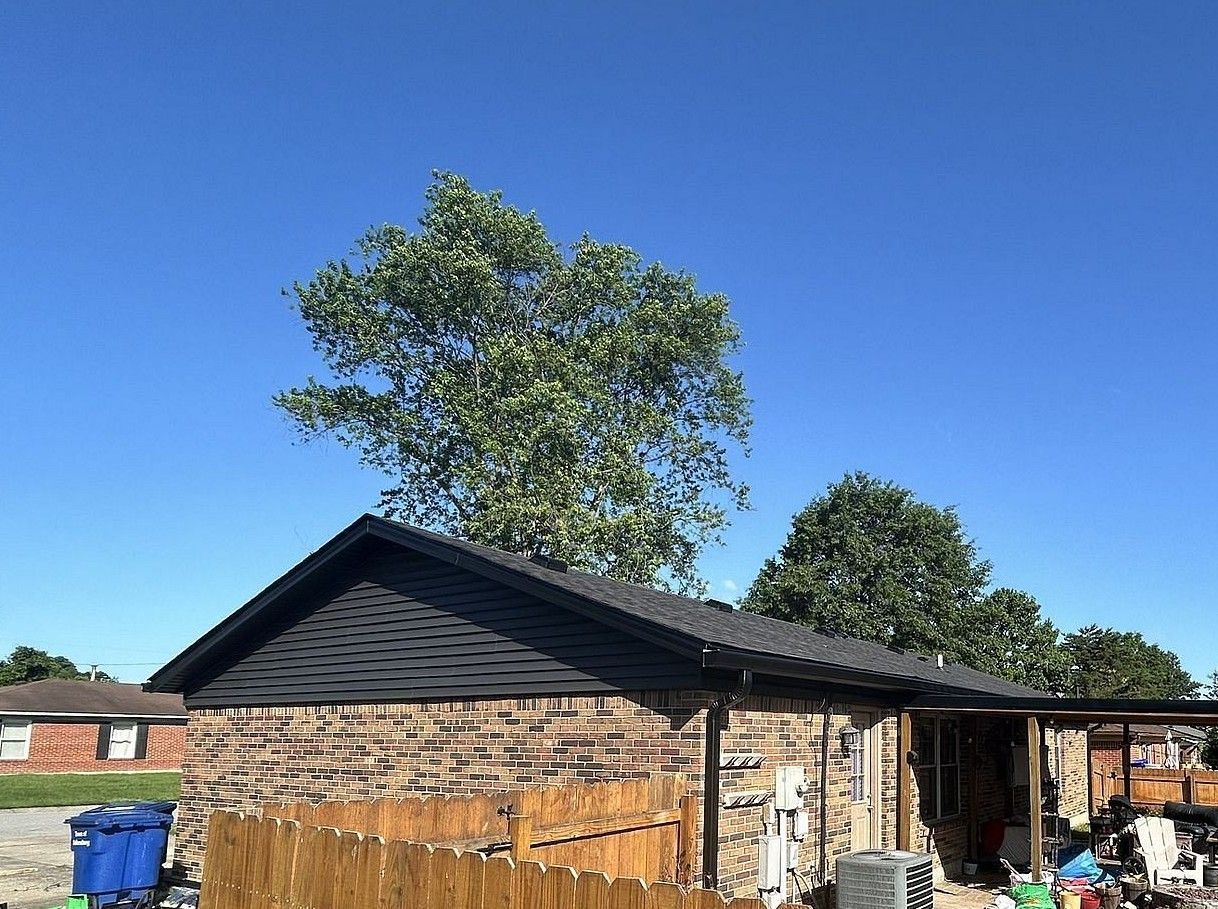 A small brick house with a black roof and a tree in the background.