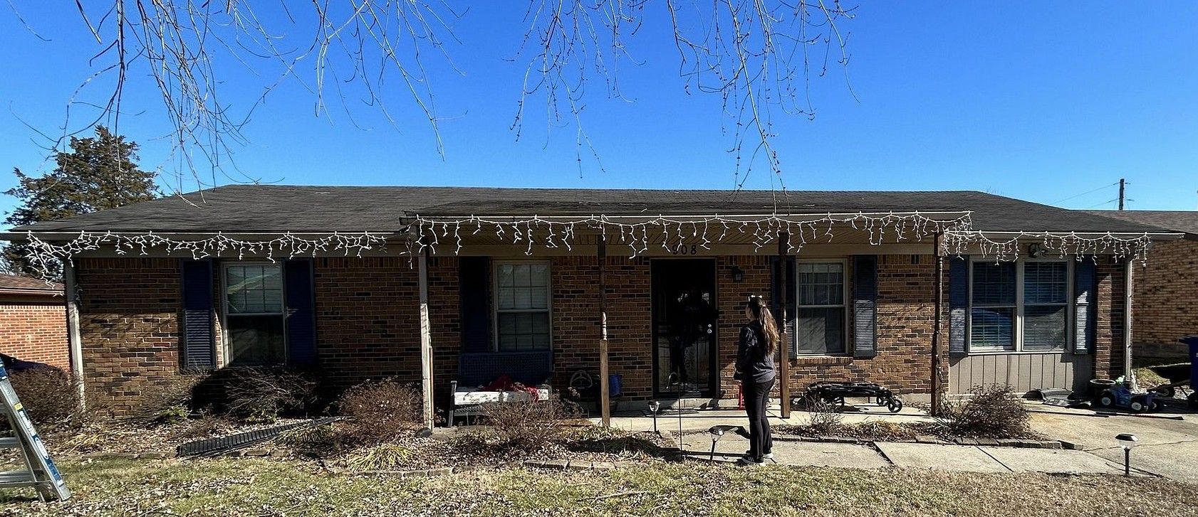 A woman is standing in front of a brick house with christmas lights on the roof.