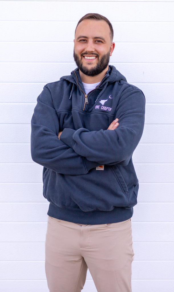 Smiling man in a navy hoodie and beige pants standing with arms crossed against a white wall