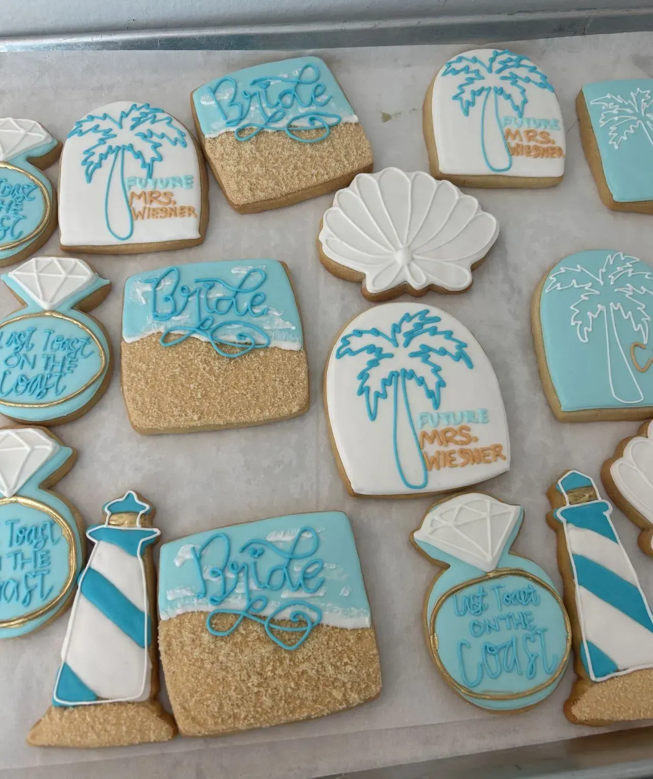 Turquoise and gold beach-themed decorated cookies on a tray, including palm trees, shells, and bridal designs.