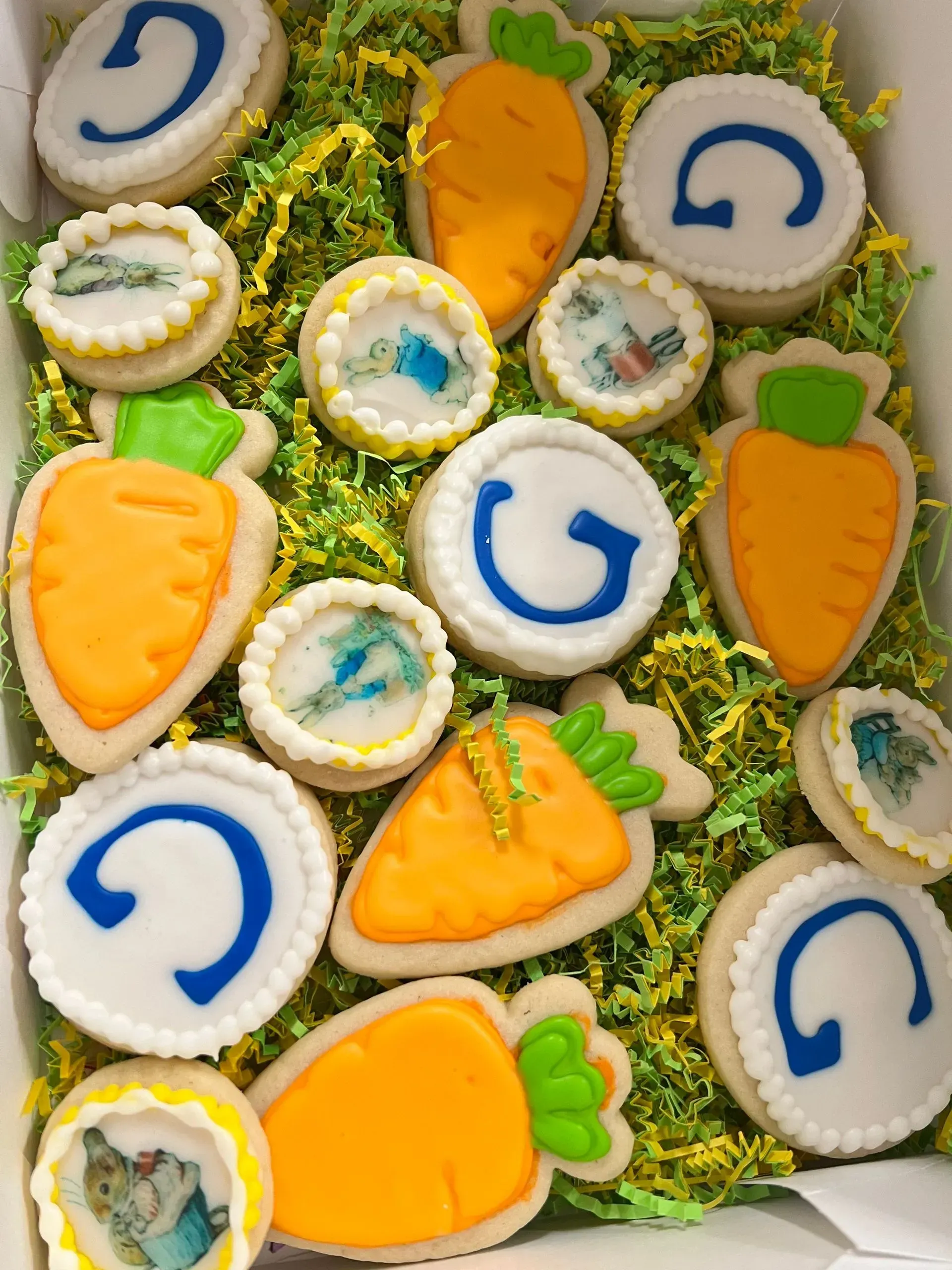 Carrot and white iced cookies arranged on green grass, some with blue letters and colorful sprinkles