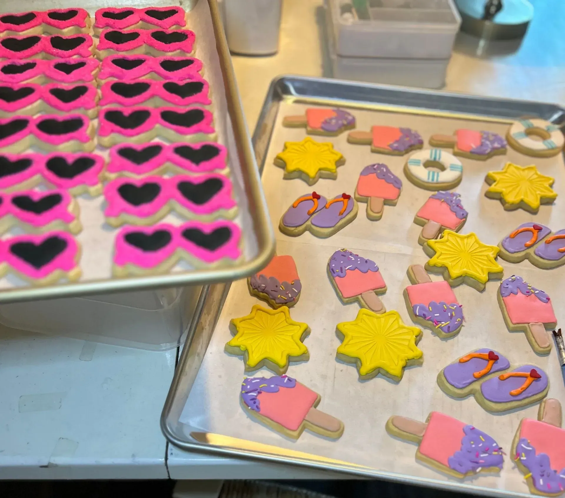 Trays of decorated cookies with pink hearts, yellow stars, and colorful popsicles on a bakery counter
