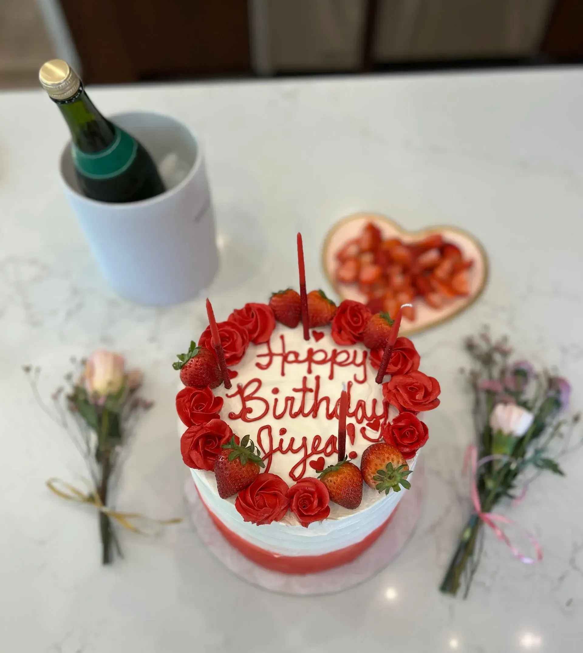 Birthday cake with red frosting and strawberries, surrounded by flowers and a bottle of champagne on a table