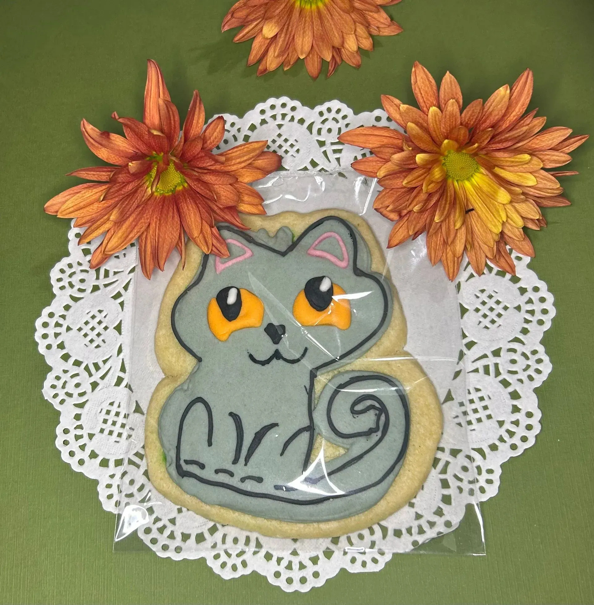 Gray cat cookie with orange eyes on a lace doily, topped with orange flowers