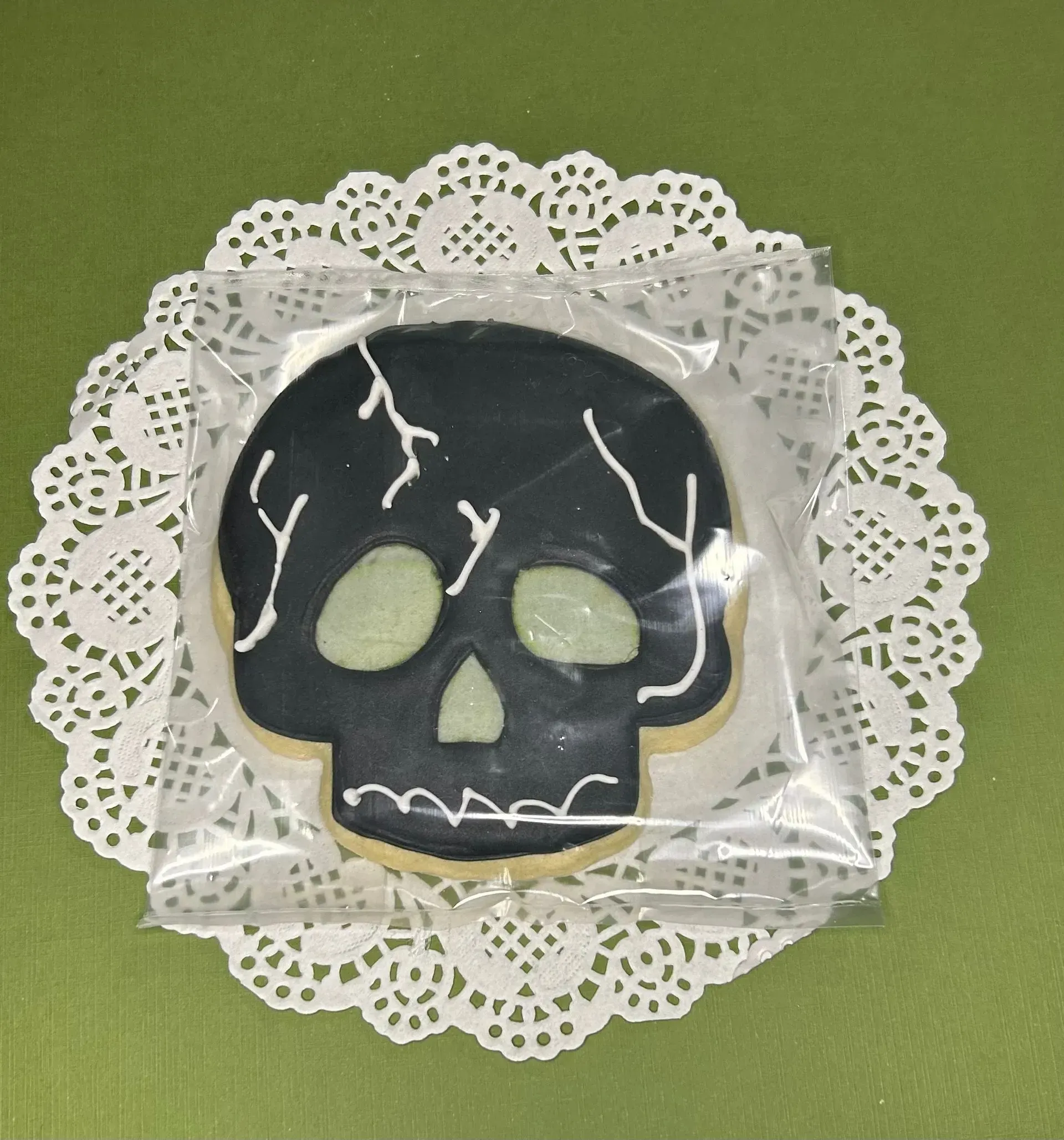 Black skull-shaped cookie with white icing cracks on a white lace doily over green background