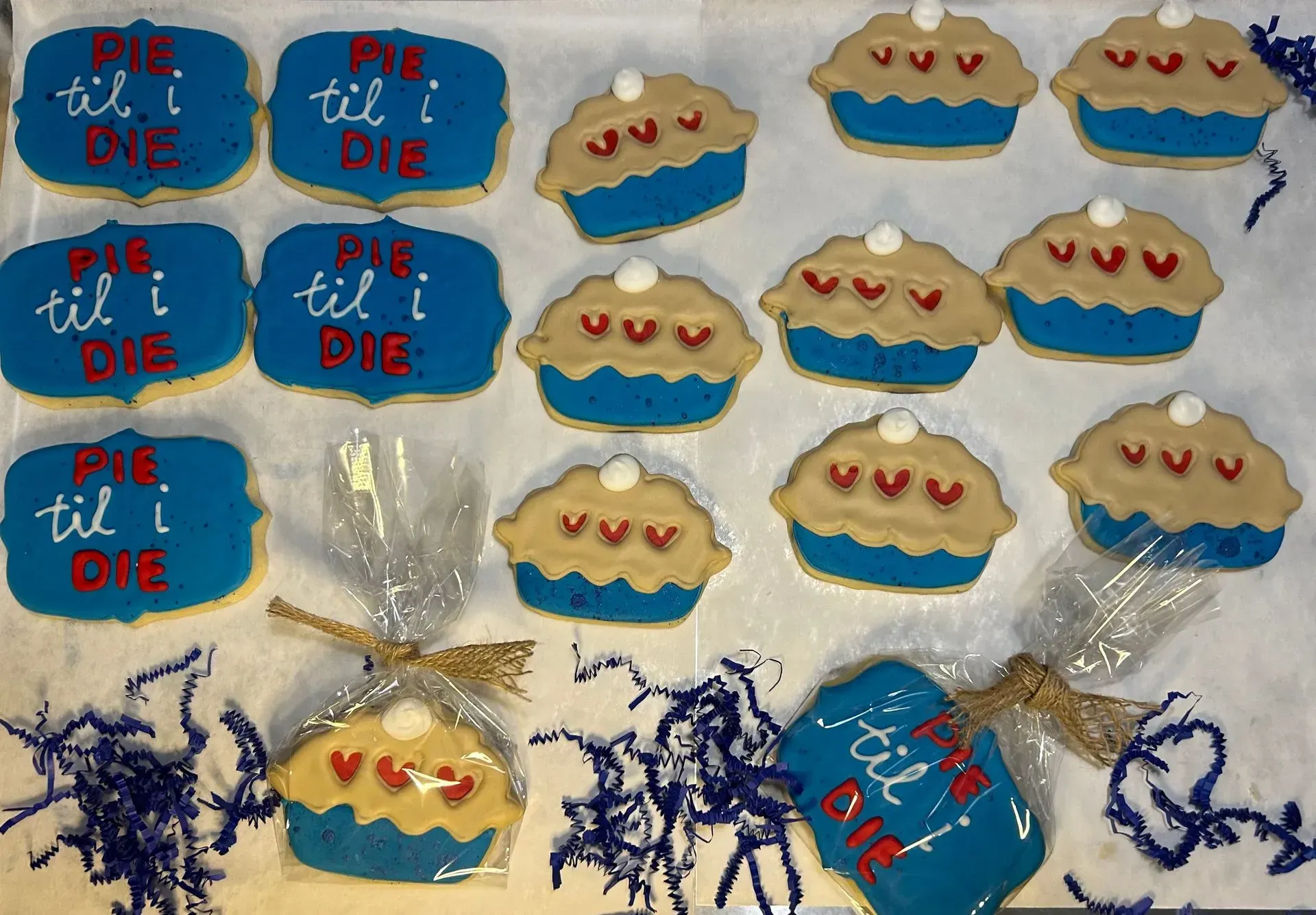 Blue and beige pie-shaped cookies and treats tied with ribbon on a tabletop, some with “PIE TIL I DIE” text.