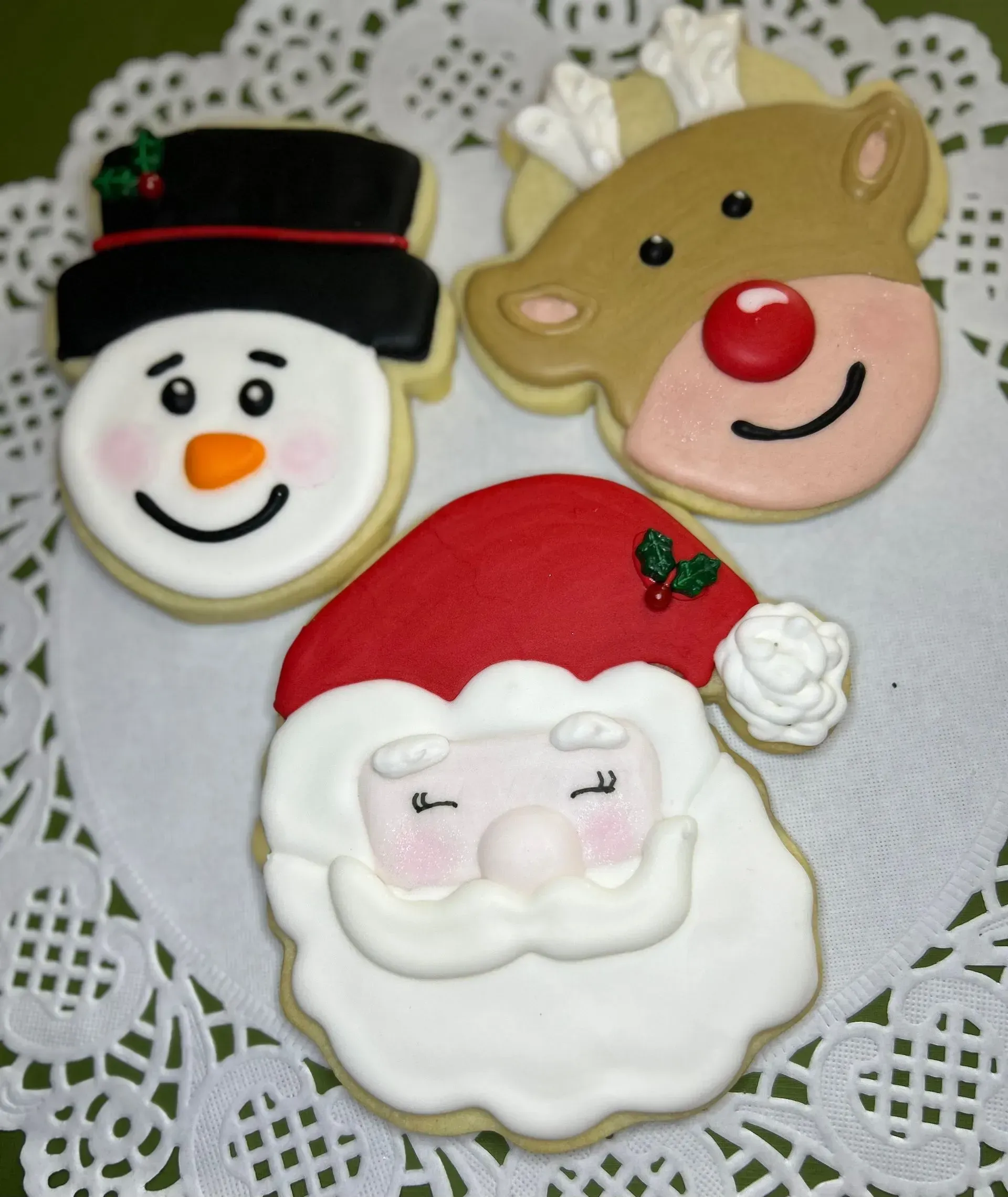 Three festive cookie faces: snowman, reindeer, and Santa on a lace doily.
