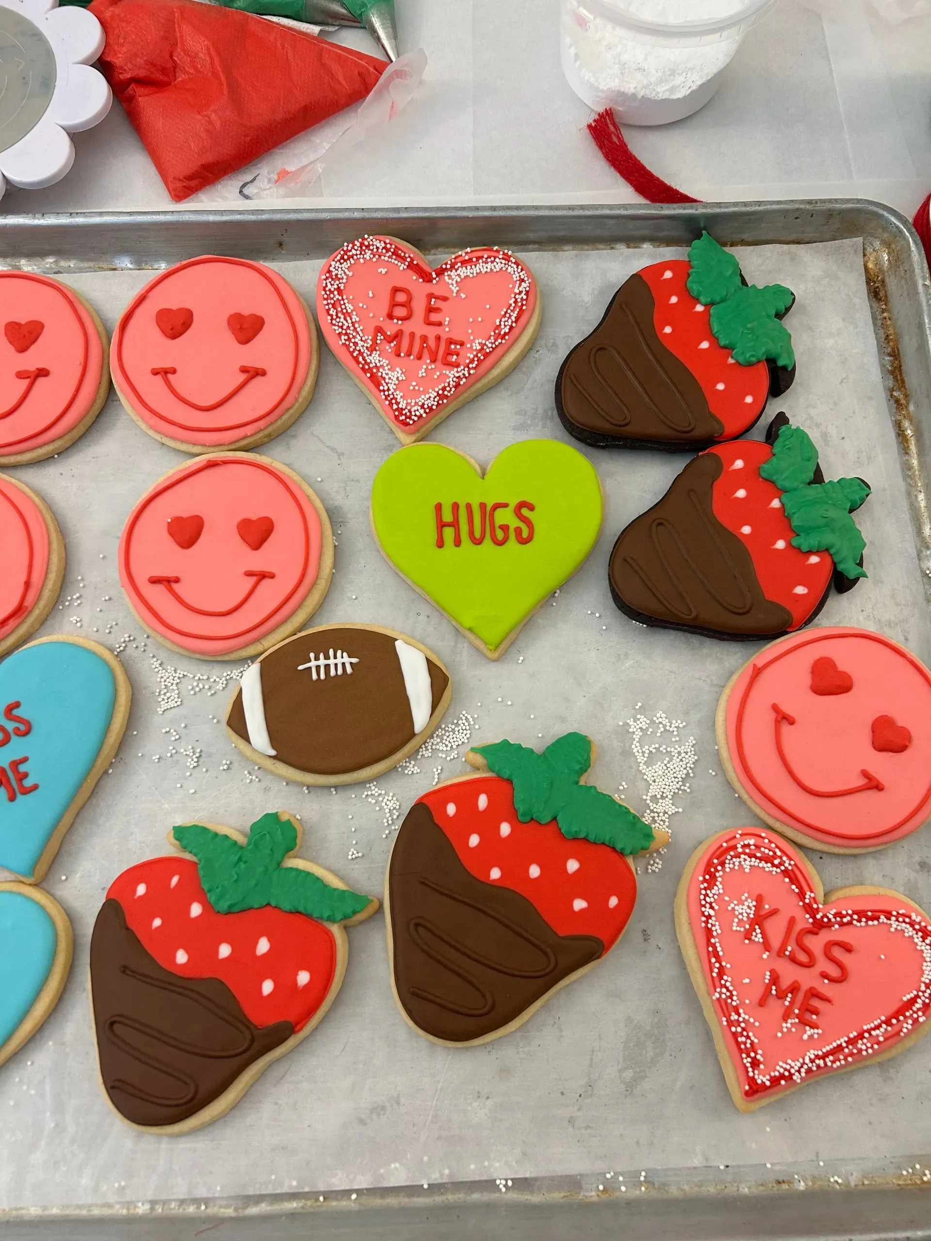 Heart-shaped Valentine cookies with pink smiley faces, chocolate strawberries, and “Hugs” and “Kiss” icing on a tray