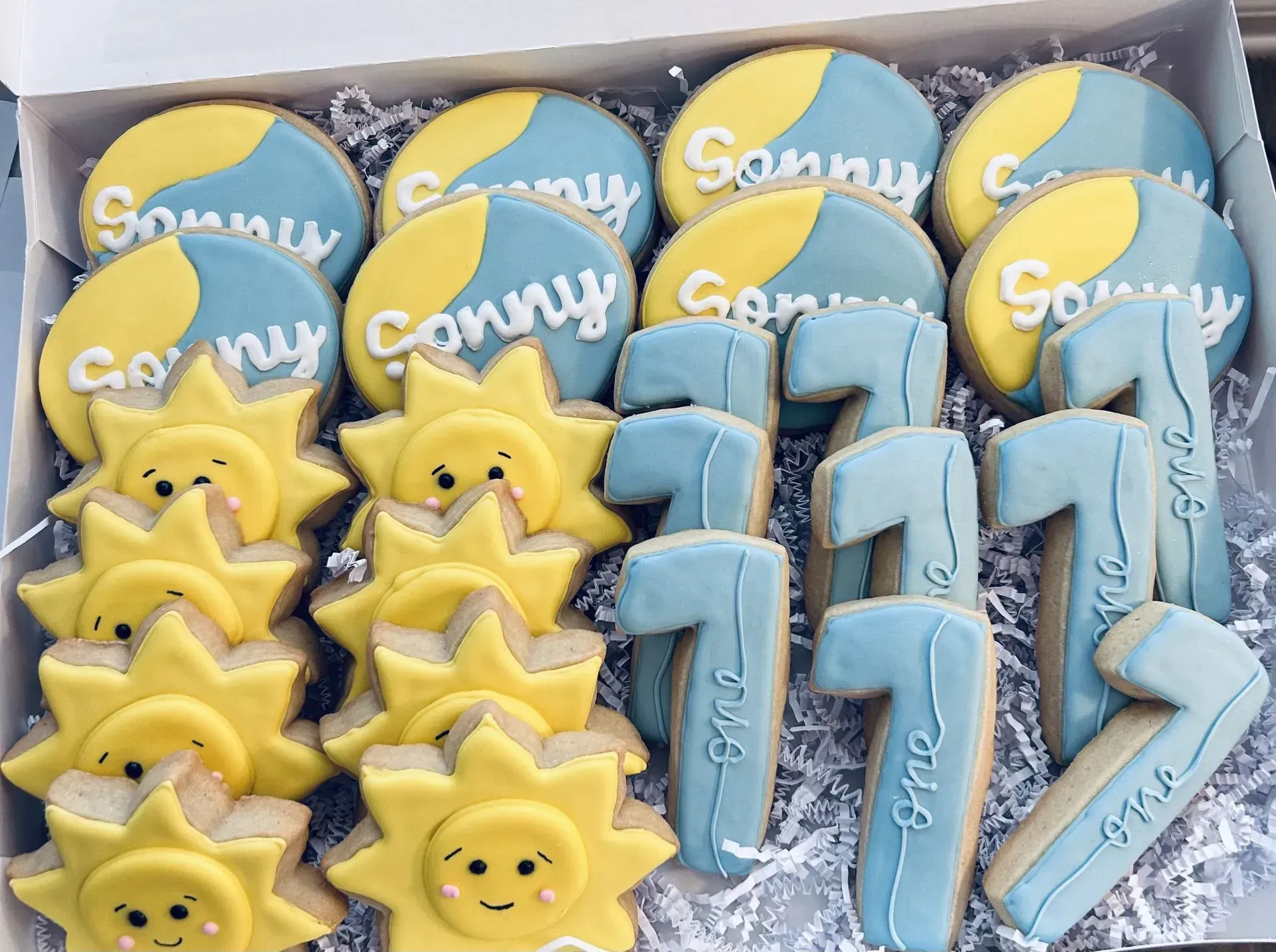 Blue and yellow decorated cookies in a box, including suns, stars, and moon shapes with name initials.