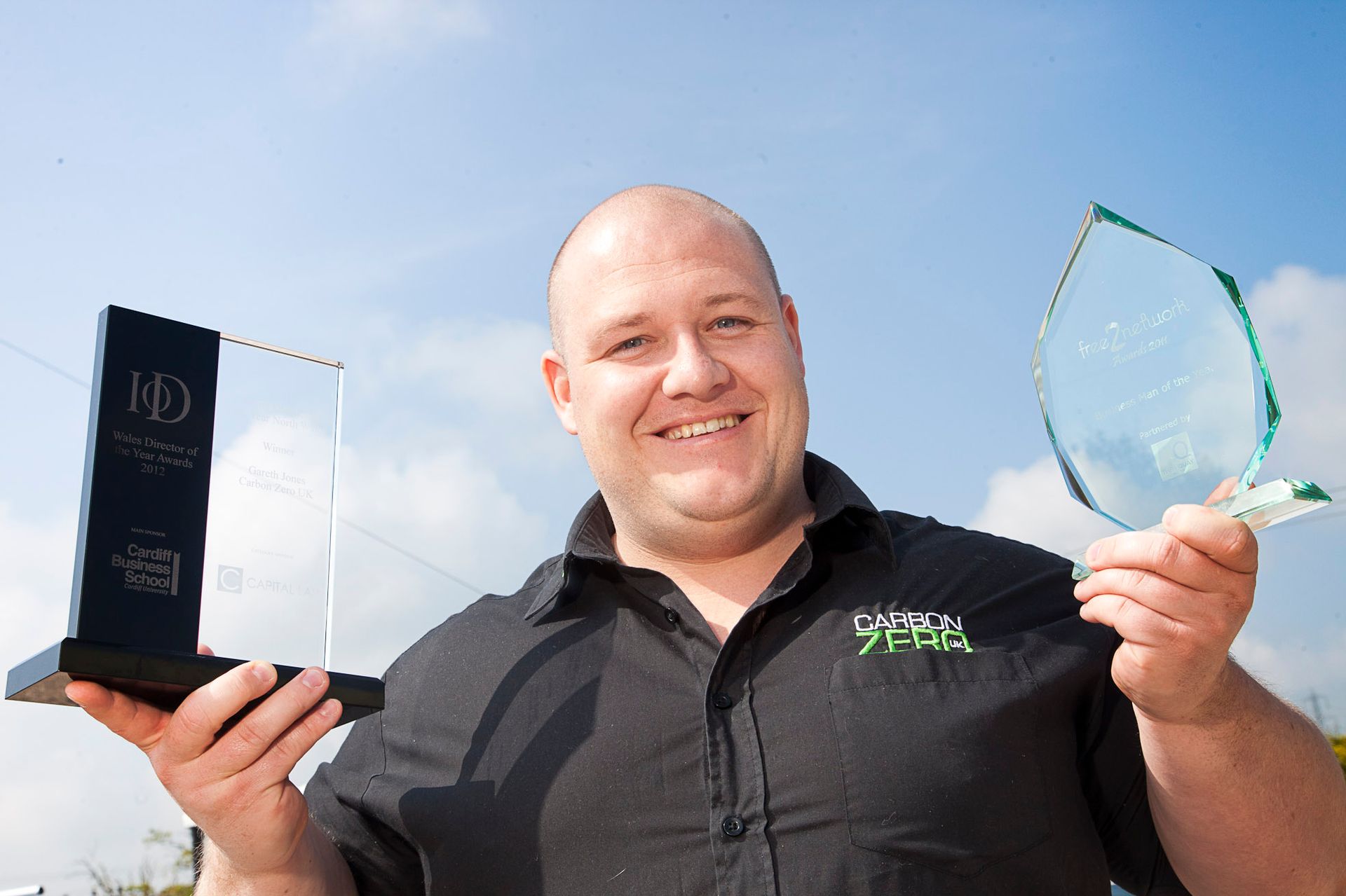 Smiling man holding an award plaque and glass trophy outdoors against a blue sky
