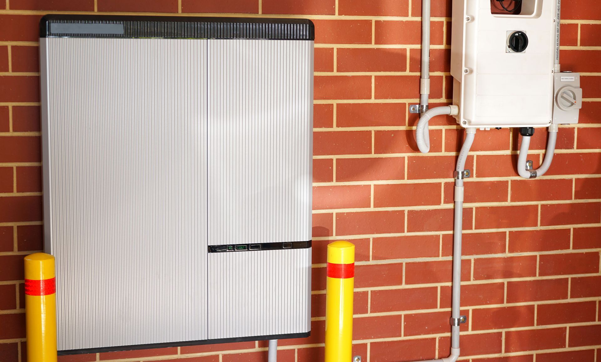 White metal enclosure beside a brick wall and electrical box, with yellow bollards in front.