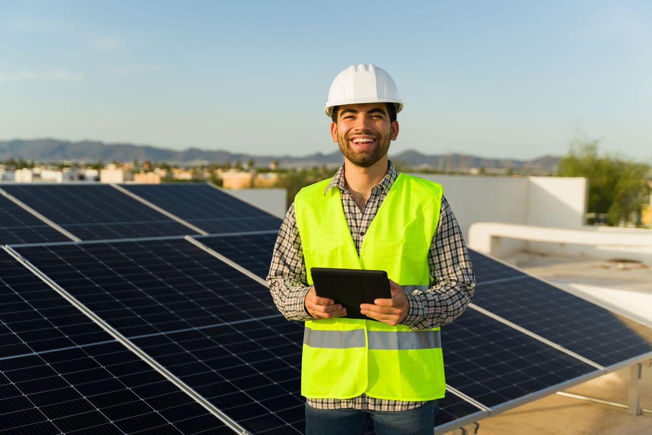 Worker in safety vest and hard hat holding a tablet beside rooftop solar panels.