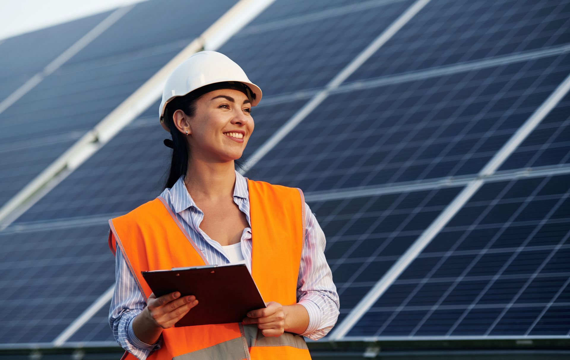 Worker in orange safety vest and hard hat holding a clipboard in front of solar panels