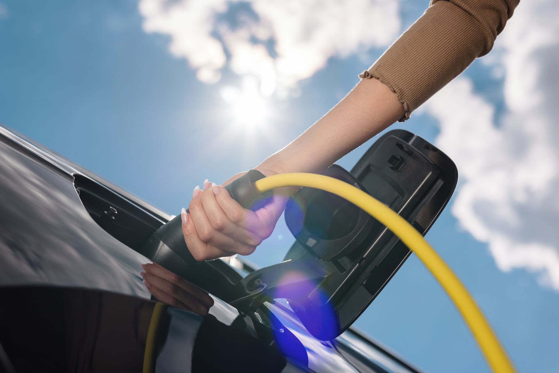 Hand plugging a yellow charging cable into a black electric car outdoors under a blue sky
