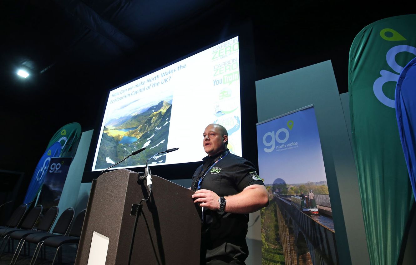 Speaker at a podium on stage beside a large projection screen and green banners at a conference