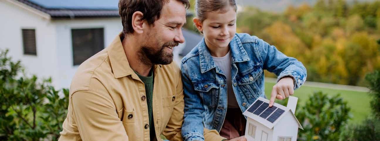 Man and child smiling while holding a solar panel model outdoors near a house