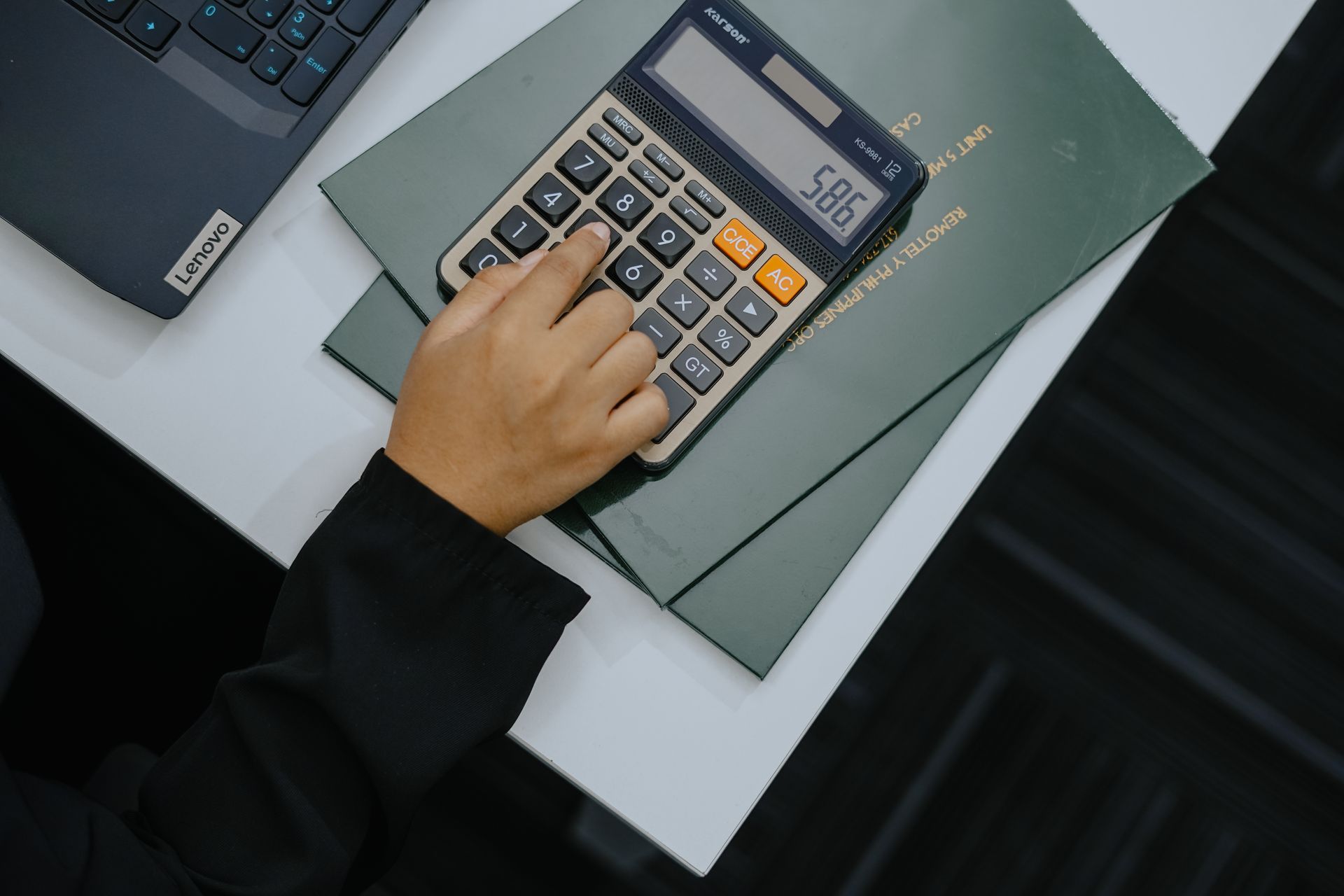 Hand using a calculator on top of documents next to a laptop on a white table.