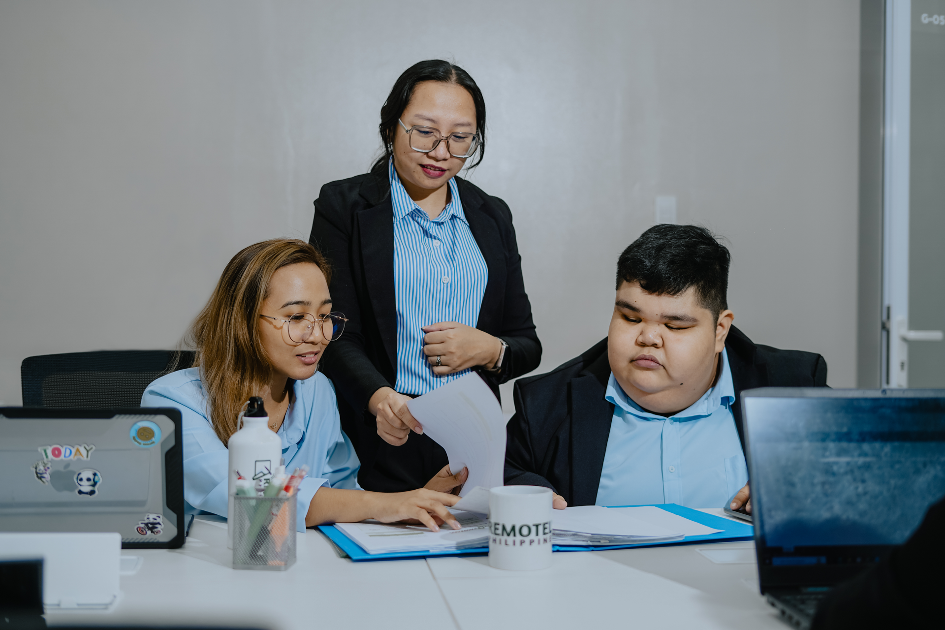 Three professionals in blue shirts and black blazers reviewing documents together at a table in an office.