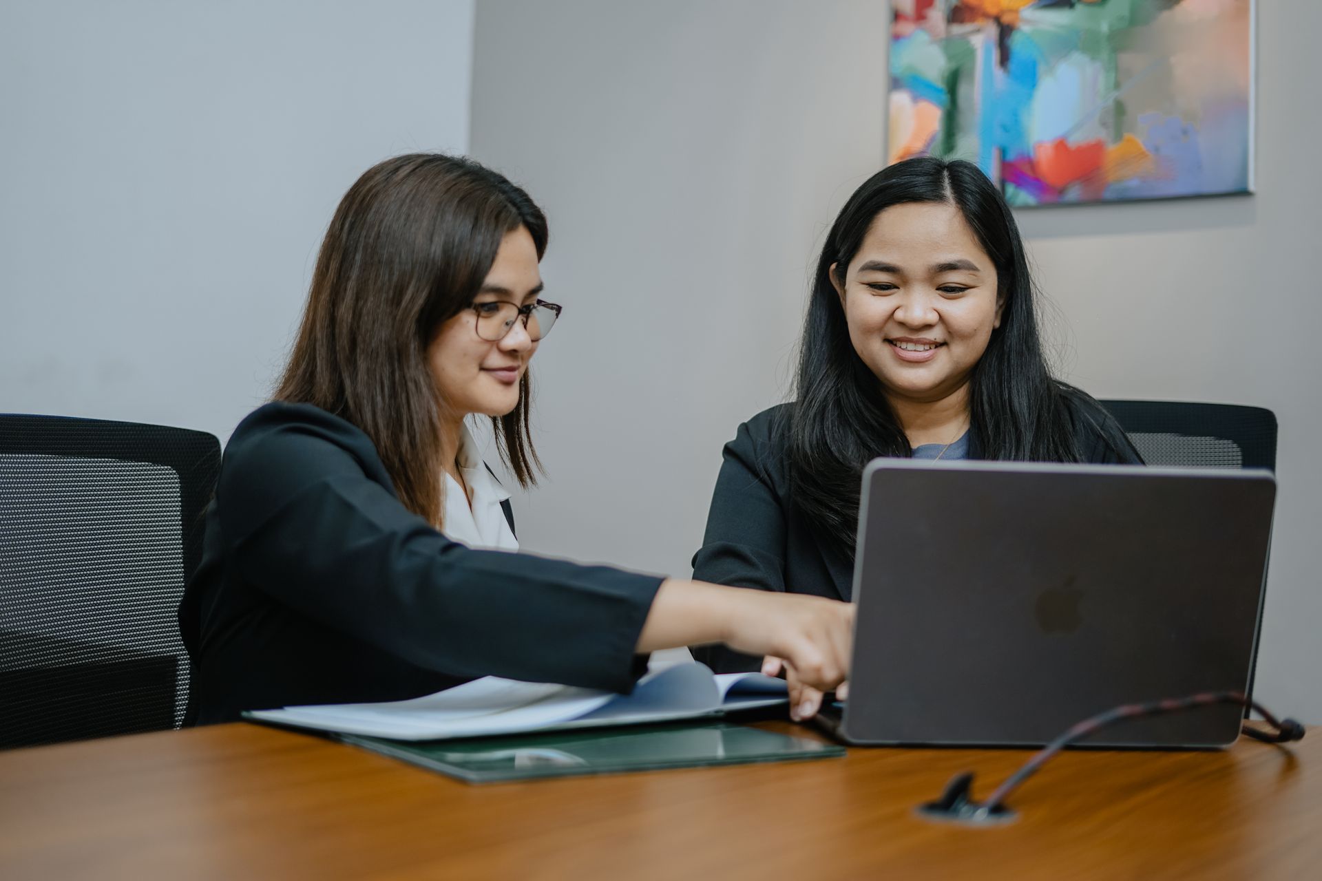 Two colleagues in business attire work together at a wooden office desk, reviewing information on a laptop and documents.