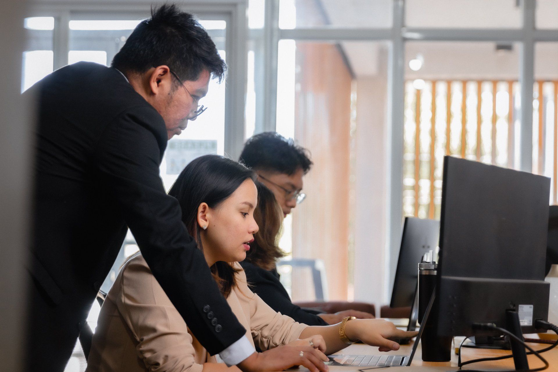 A man in a black blazer leans over a woman’s desk as they look at a computer screen together in a bright office.