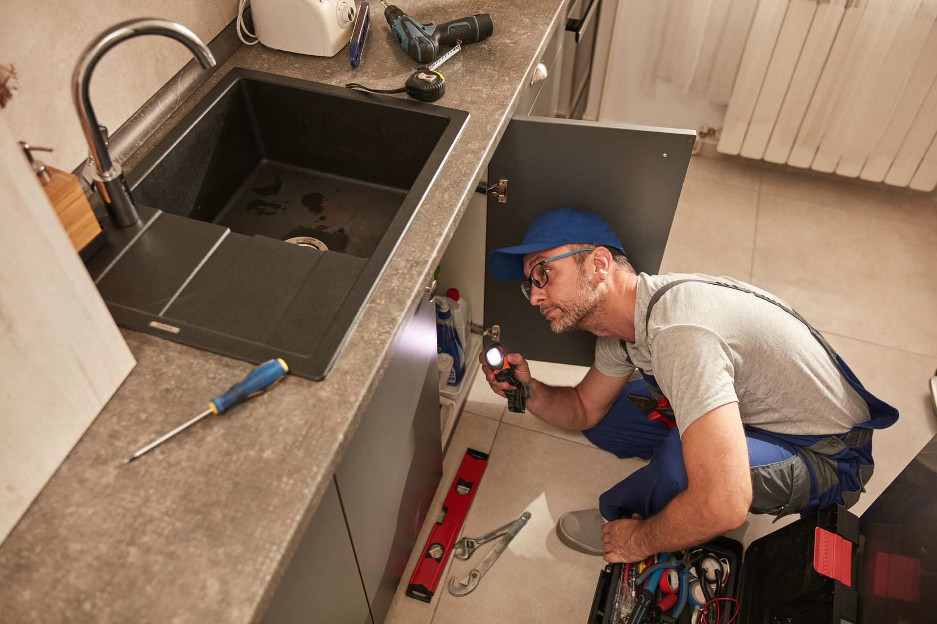A plumber fixing and repairing water pipes under a kitchen sink.