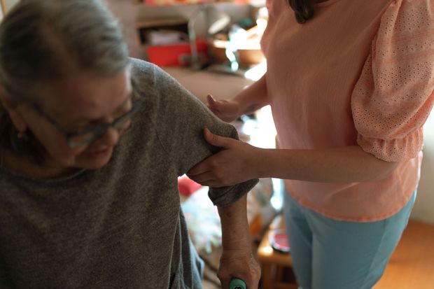 Person helps an older adult walk indoors, holding their arm.