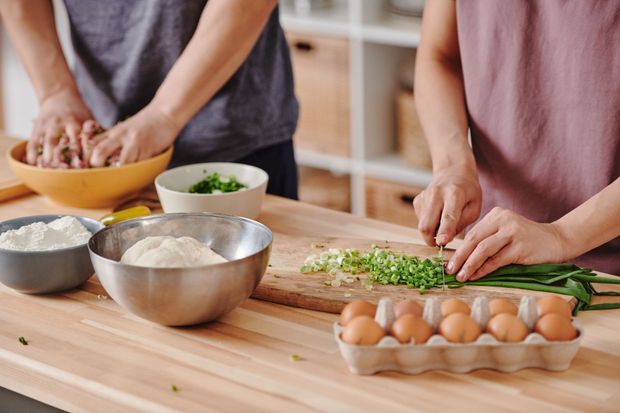 Two people preparing food at a wooden table with eggs, flour, and chopped herbs in a kitchen