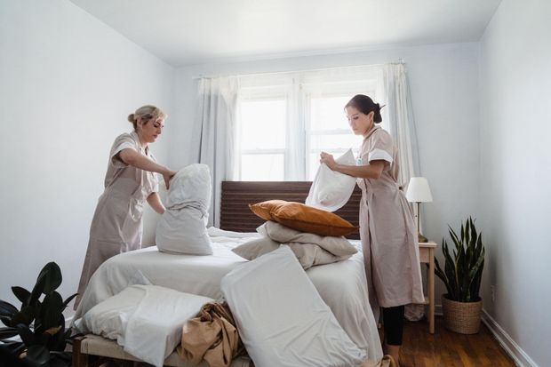 Two people making a bed in a bright bedroom with stacked pillows and blankets