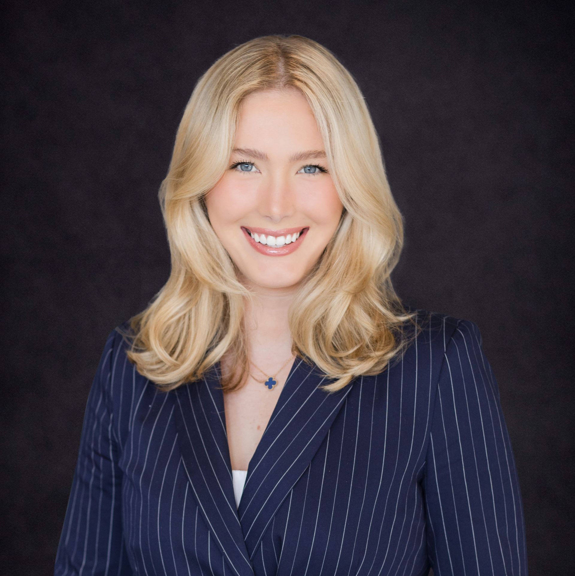 Smiling blonde woman in a navy pinstripe blazer against a dark background
