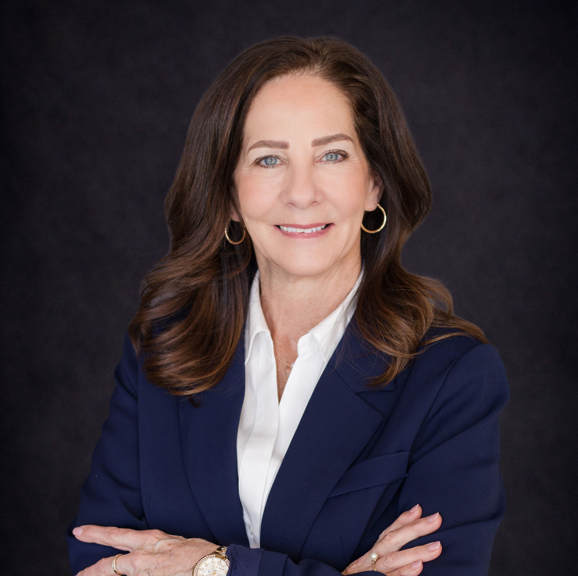 Professional headshot of a woman in a navy blazer and white blouse, smiling with arms crossed on a dark background