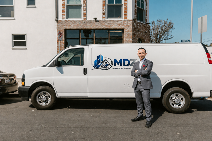 Man standing beside a white MDZ service van in front of a building, wearing a gray suit and red tie