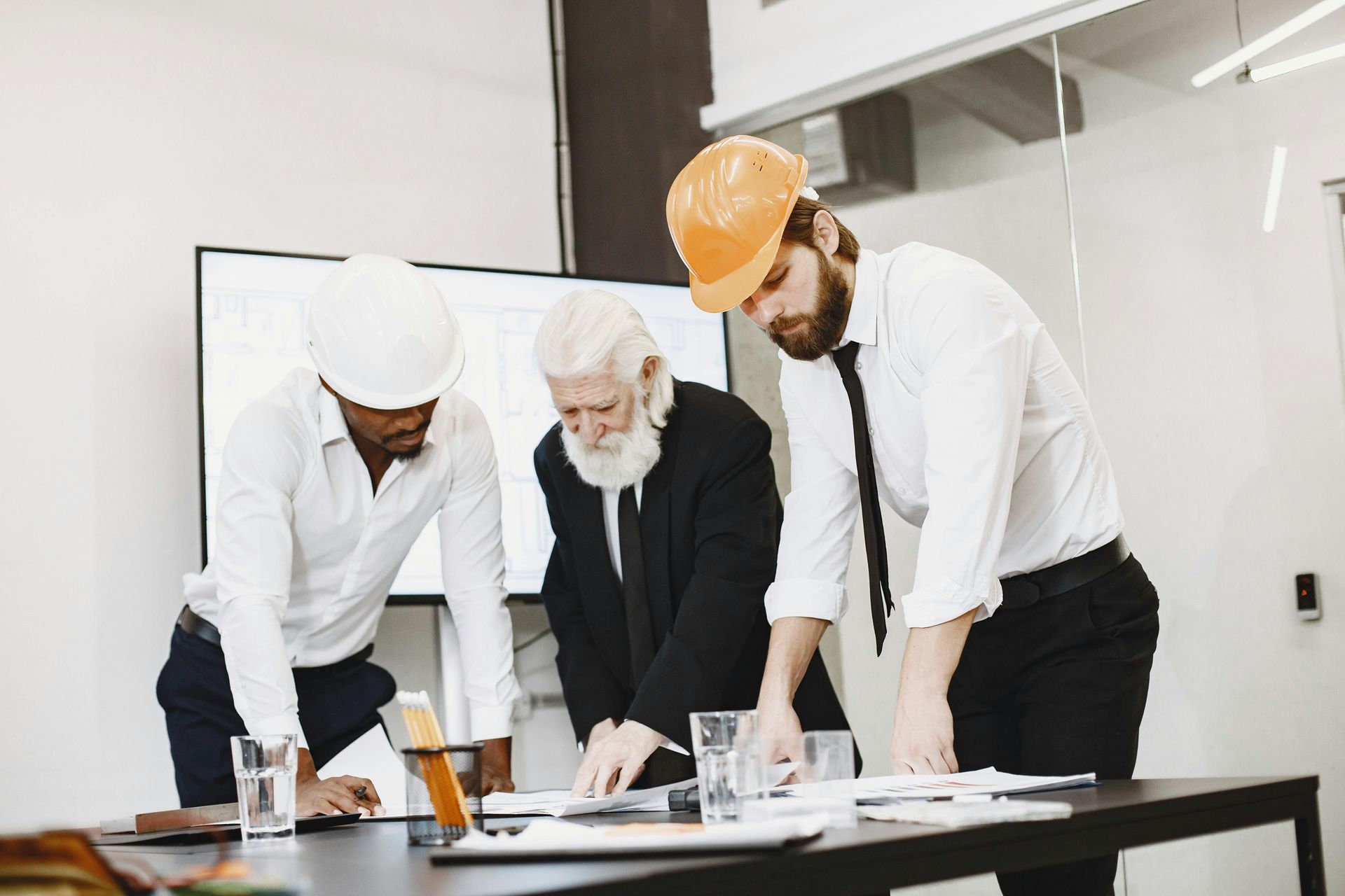 Three workers in hard hats reviewing plans at a table in an office or construction meeting room