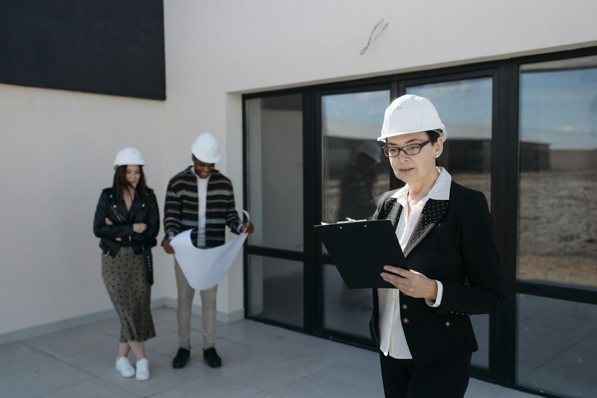 Three people in hard hats review plans outside a modern building.