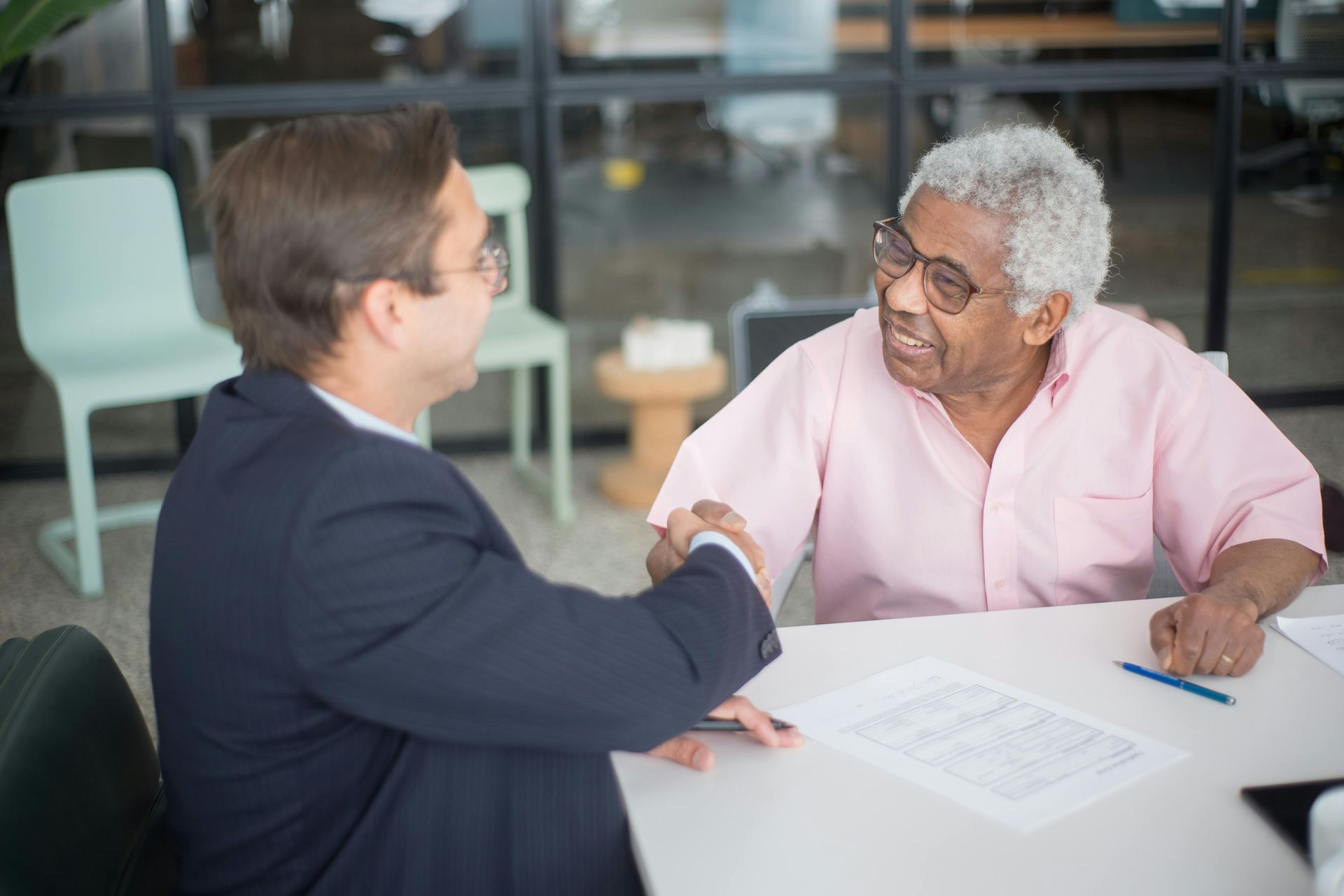 Two people in a meeting shake hands across a table in an office setting.