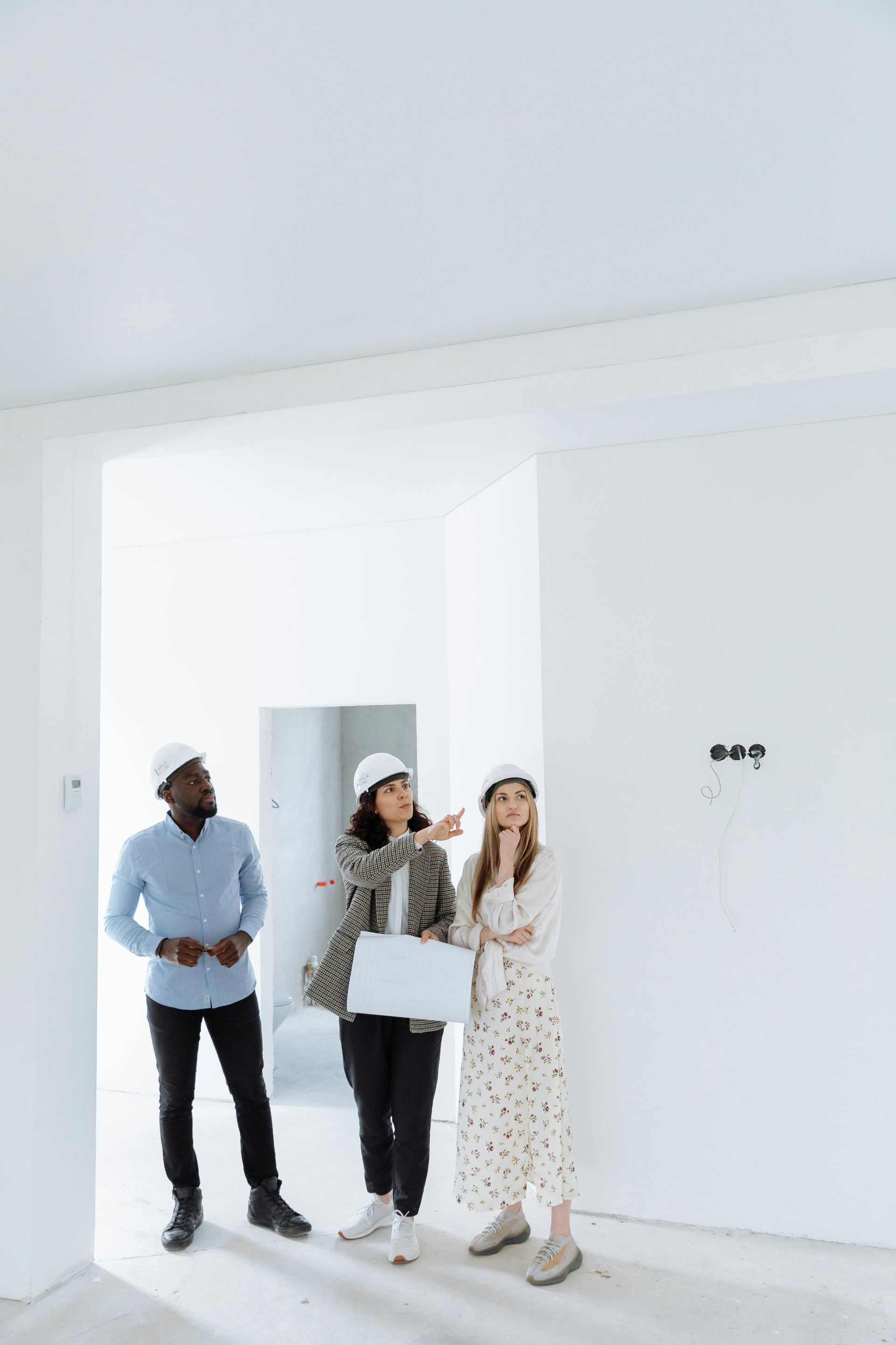 Three people standing in a bright, empty room, looking at the walls and windows.