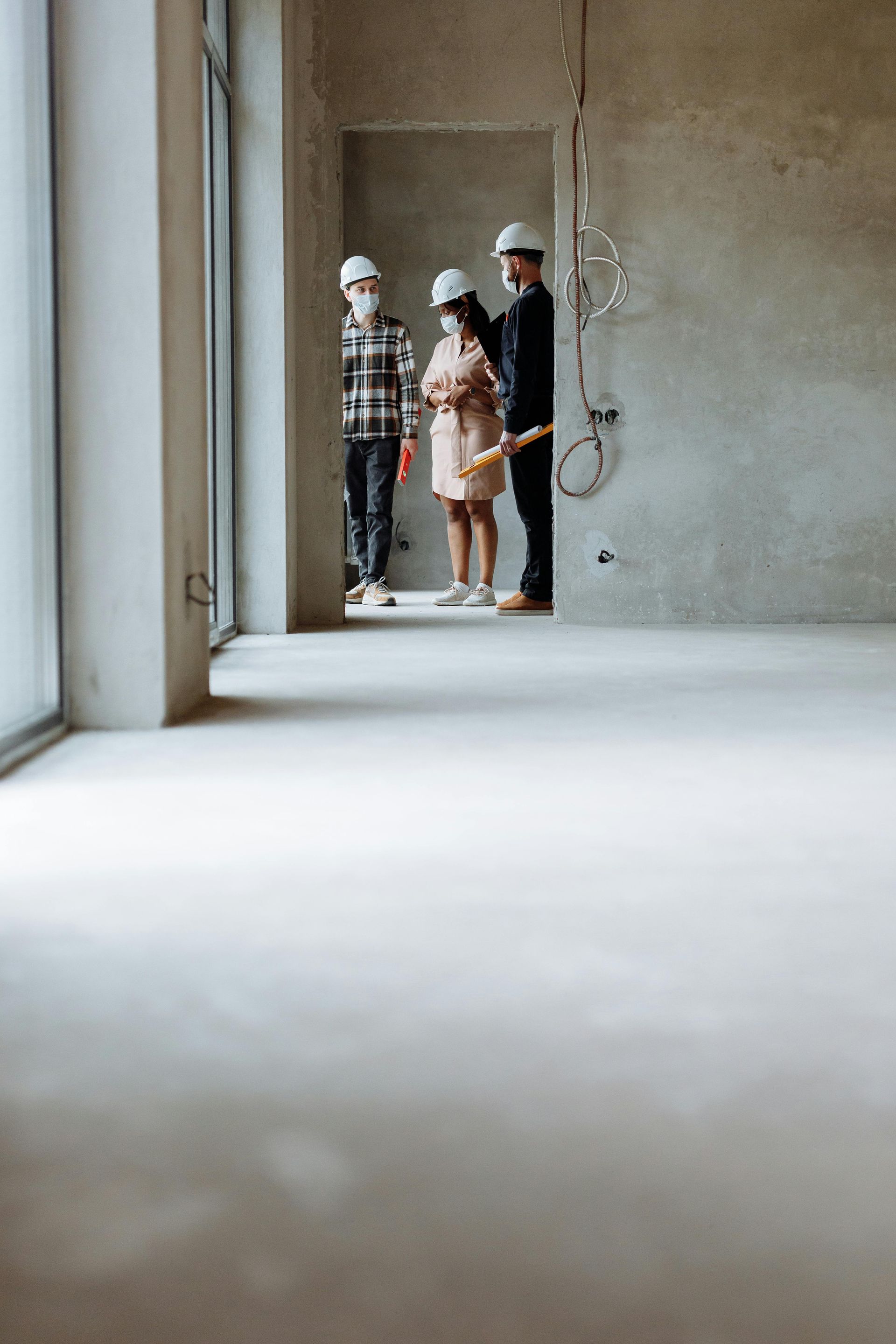 Three people in hard hats stand in an unfinished concrete room near a window.