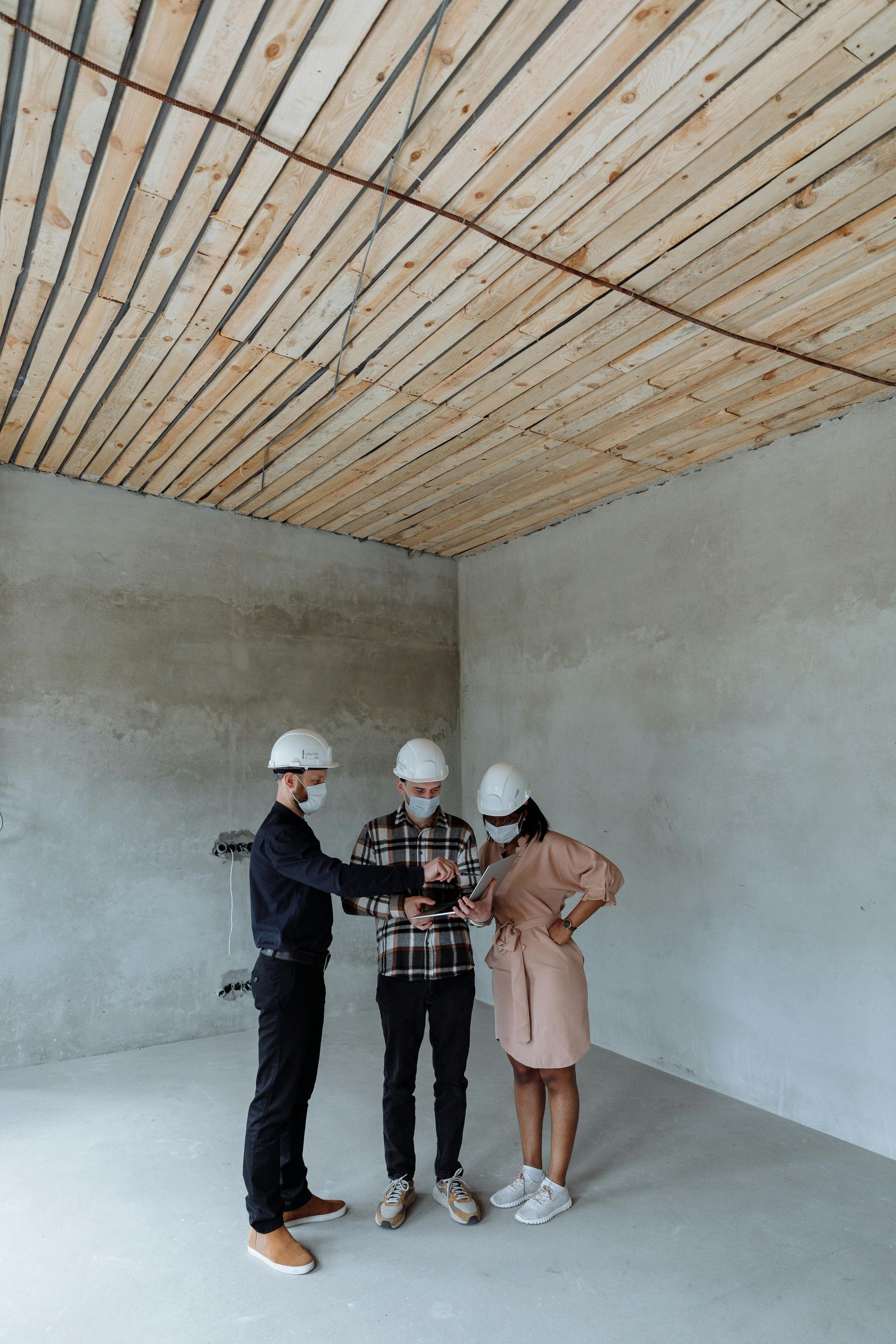 Three people in hard hats standing in an unfinished room with concrete walls and a wood-slat ceiling.