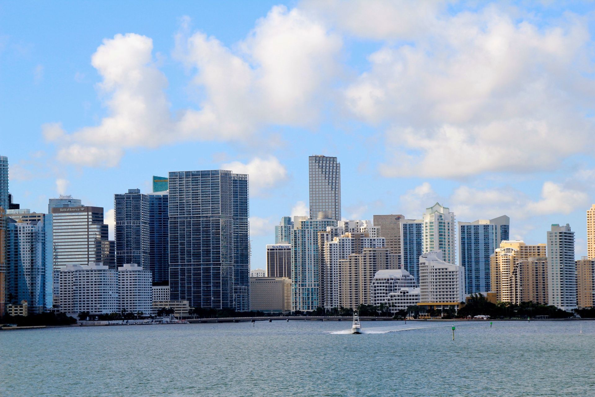 City skyline of modern waterfront buildings under a cloudy blue sky, with a sailboat on the water.