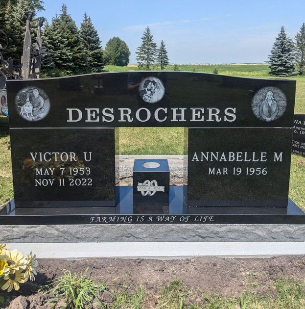 Black granite Desrochers gravestone with white lettering in a sunny cemetery, flanked by trees and flowers